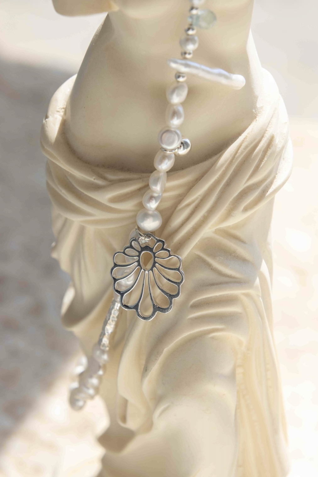 A close-up of a silver-toned beaded necklace with a flower-shaped pendant and pearl-like beads draped over a cream-colored draped statue against a soft, bright background.