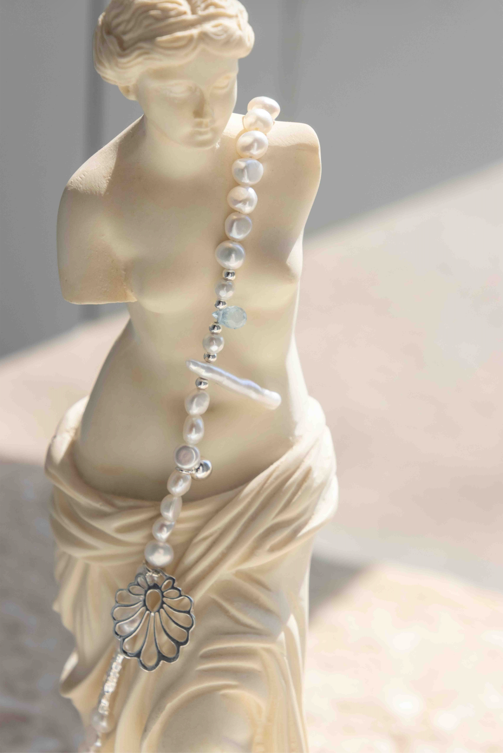 A close-up of a draped classical-style statue in warm ivory, with a pearl-like beaded necklace draped over its torso and a silver flower pendant hanging near the lower draped fabric.