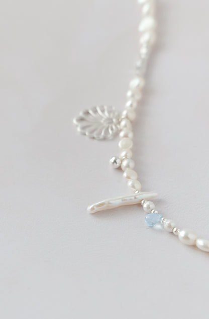 A delicate silver necklace with white pearl-like beads, a silver filigree heart charm, a small silver cross charm, and a tiny light blue faceted bead, shown on a light gray background.