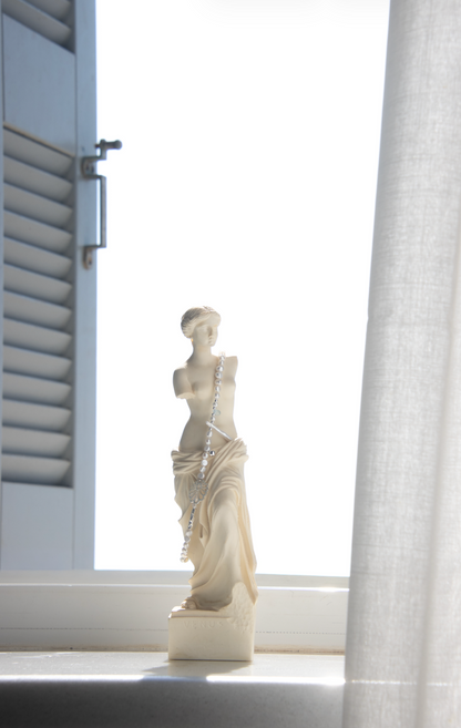 A small cream-colored classical female statue with a silver chain draped around the waist, displayed on a windowsill with bright natural light, a white curtain, and window shutters in the background.