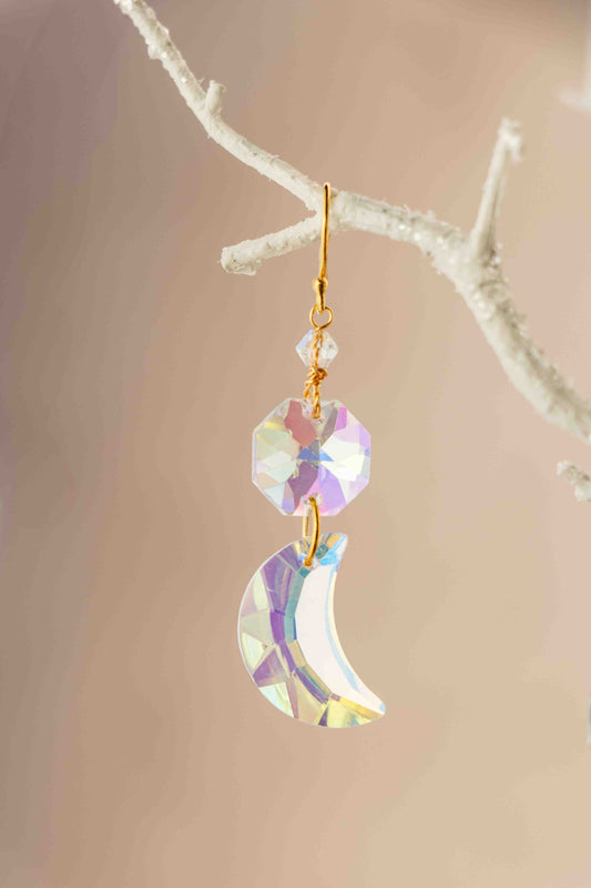 Iridescent moon-shaped earring with gold accents on a branch against a beige background