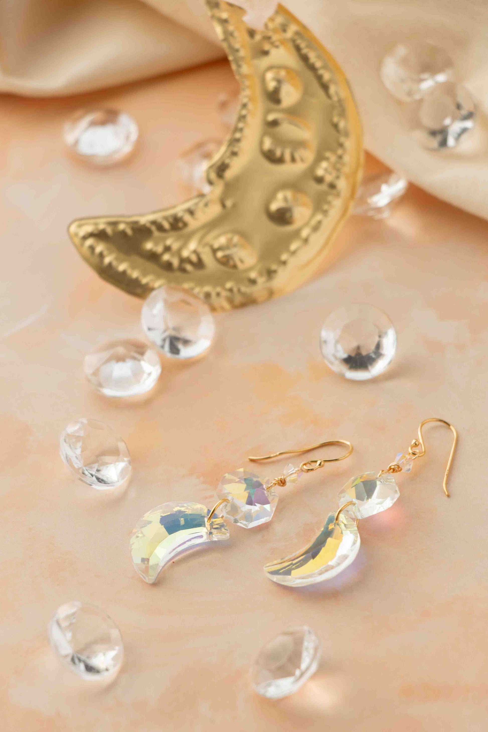 Gold moon-shaped earrings on a soft beige background with clear crystals.