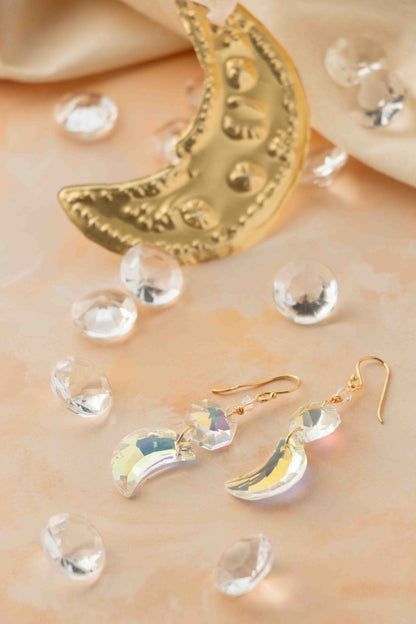 Gold moon-shaped earrings on a soft beige background with clear crystals.
